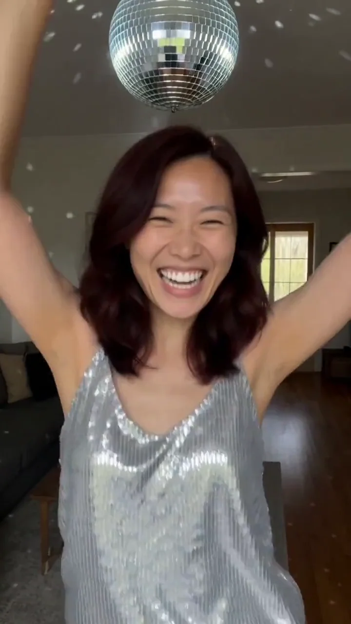 AI generated fictional adult woman in a silver sequin top dancing with arms raised under a disco ball